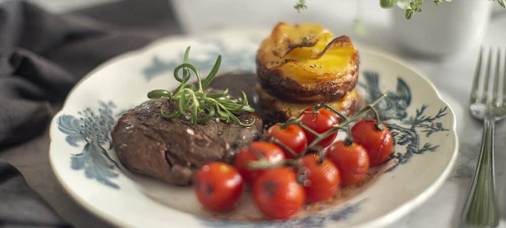 Oxfilé med grillade tomater och gratäng på tallrik.