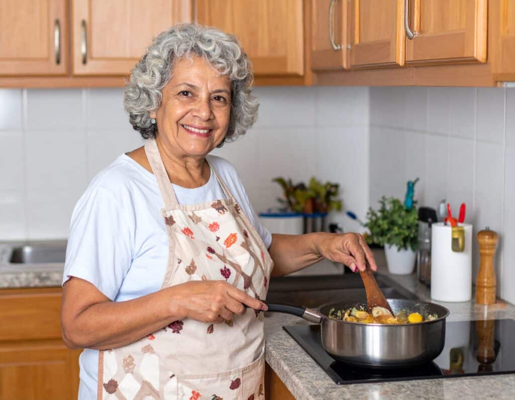 Äldre kvinna lagar mat i kök och rör om i stekpanna.