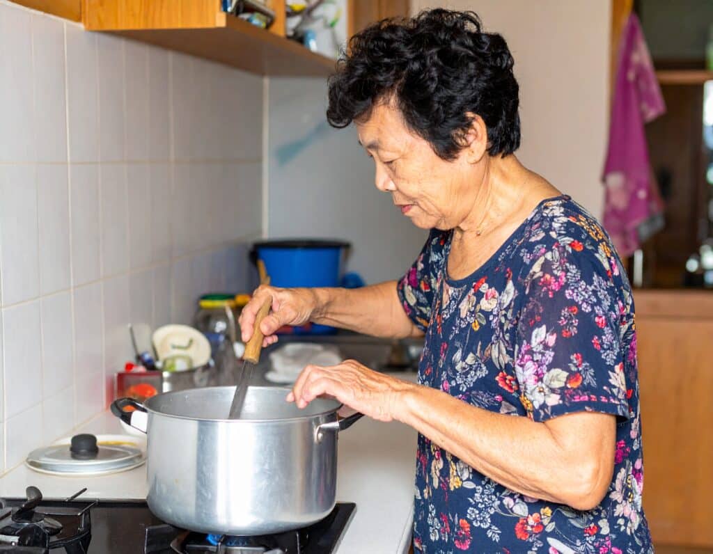 Äldre kvinna lagar mat och rör i gryta vid kökspis.