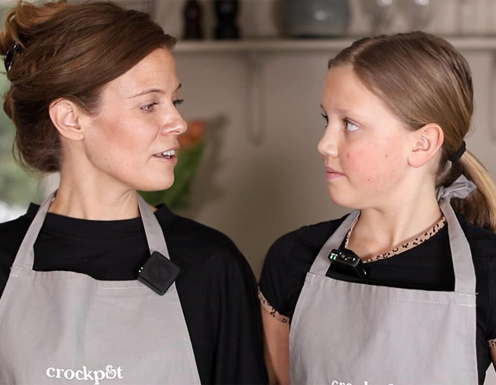 Mamma och dotter i köket, klädda i förkläden, pratar och lagar mat.